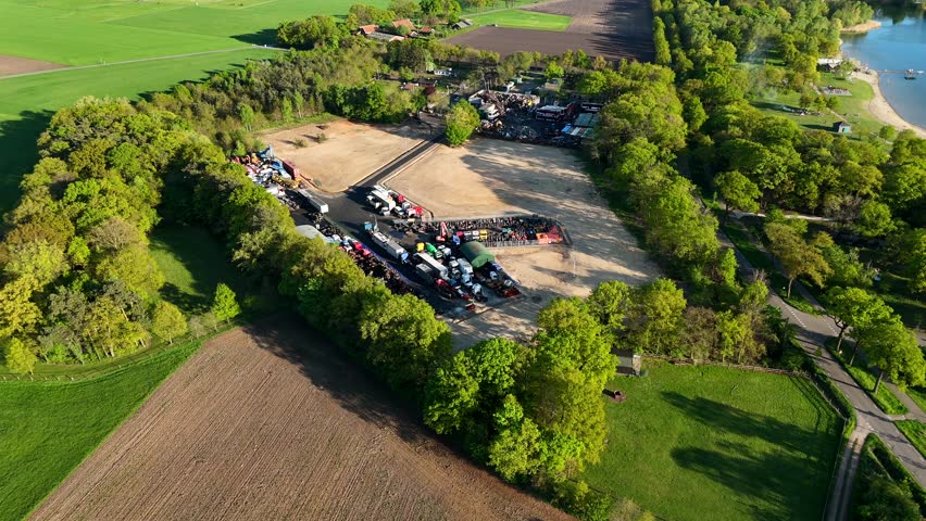 Industrial junkyard field in rural american countryside near lake. Sunset time in usa. Aerial top down flyover. Green trees lighting in the evening. Parking destroyed cars and trucks in suburb.
