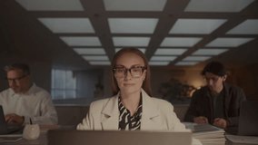 Young confident businesswoman using laptop at desk in modern open-plan office space with two male coworkers working in background during evening - Powered by Shutterstock - Get 15% off with code: PIKWIZARD15