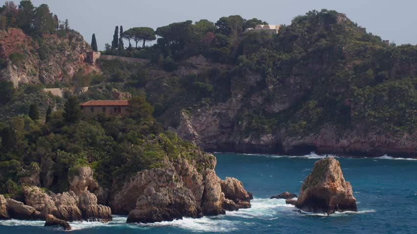 Close-up of Isola Bella’s back side showing a house and blue ocean waves hitting rocks, in Taormina, Sicily, Italy (Italia)