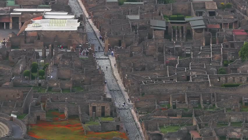 Aerial view of Pompeii in Italy, showing stone streets, arched doorways, columns, and visitors walking among the ancient ruins near Mount Vesuvius.