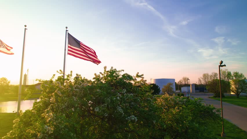 American and Ohio state flags waving at sunset beside industrial park and pond