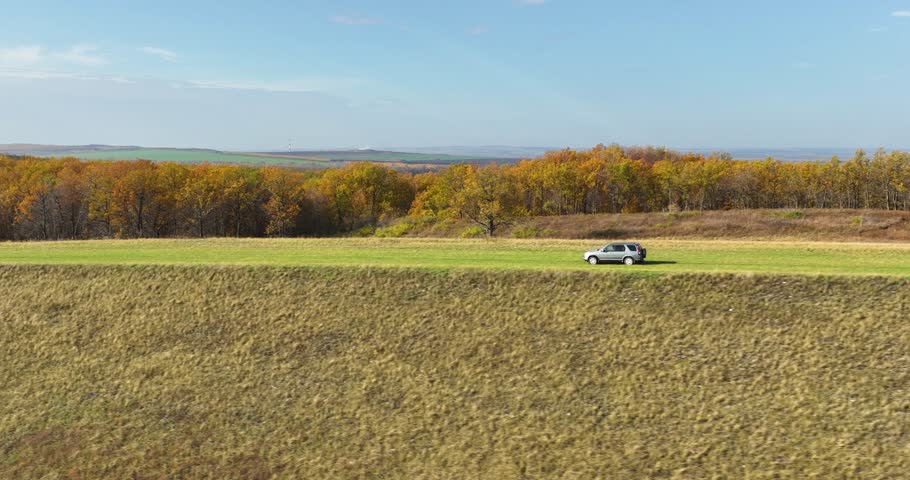 One gray crossover SUV car driving alone freeway trip, travel on gravel roathrough colorful forest background at autumn sunny sunset - Aerial drone view
