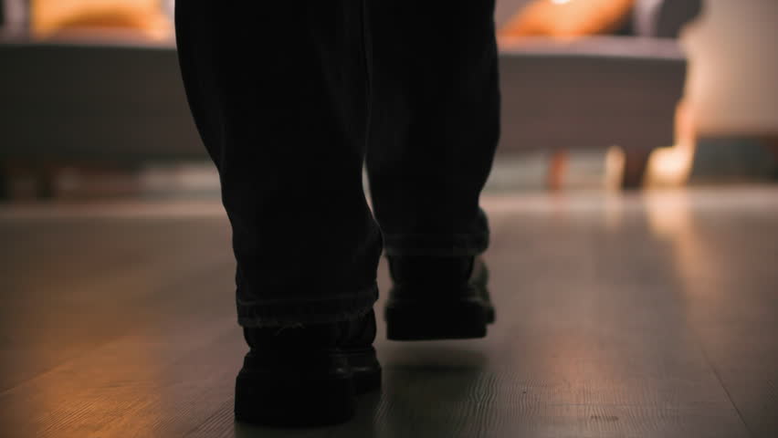 A woman in her outerwear,denim pants and boots walks across the room to the sofa. Close-up shot. Ground level view. Slow motoin