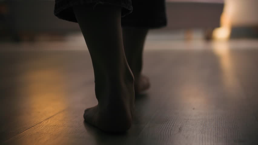 Barefoot woman in denim pants walking on wooden floor towards sofa. Close-up shot. Ground level view. Slow motoin