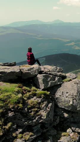 A woman sitting on top of a mountain looks into the distance at the beauty of the Carpathian mountains. Aerial view. Silhouette of a woman sitting on a rocky mountain. View of a young girl from the