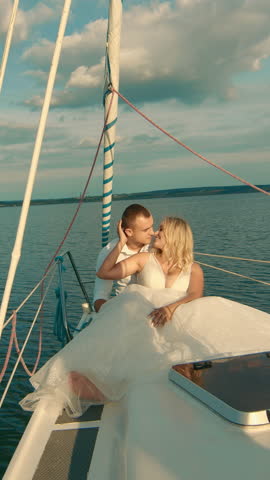 Brides sit on a yacht, looking at each other. Beautiful landscape behind them. The bride smiles tenderly at the groom