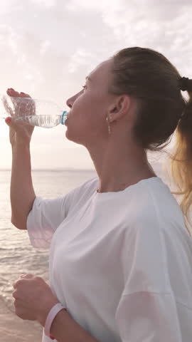 Quench thirst after morning run or workout on seashore, woman drinking water. Slender young adult lady sipping fresh water from plastic bottle, admiring sunrise or sunset above ocean, training outdoor