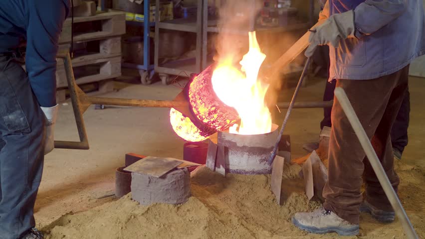 Workers pour molten bronze from crucible into mold in bell casting foundry, slow motion