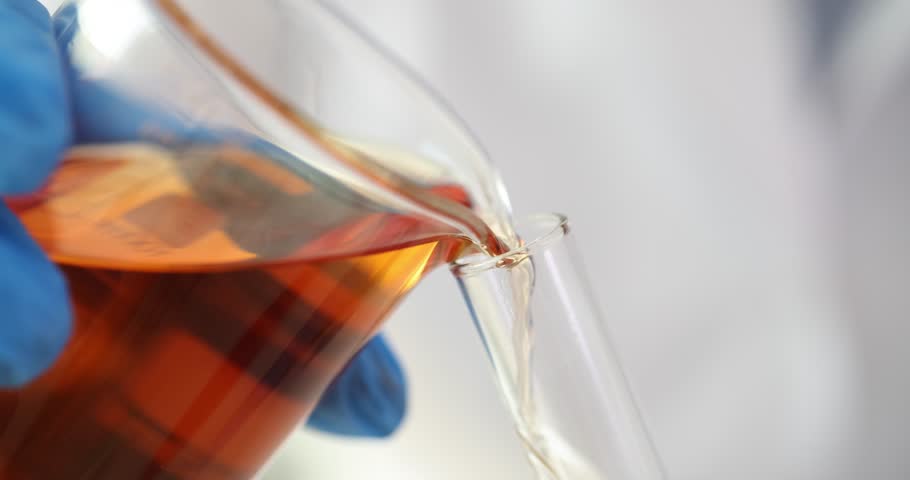 Gloved hand pours orange liquid from beaker into test tube in lab. Technician conducts scientific experiment with color fluid substance in laboratory