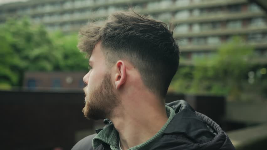 Medium close-up of young man with clean beard standing in Barbican with visible concrete housing in background, staring forward with earnest intent stare