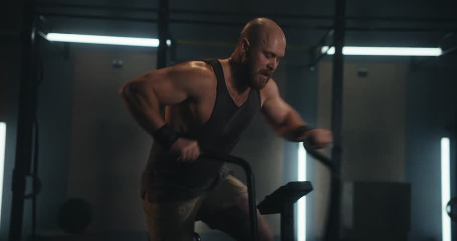 Zoom Out strong male athlete doing intense workout and fast movement while exercising on exercise bike in modern gym