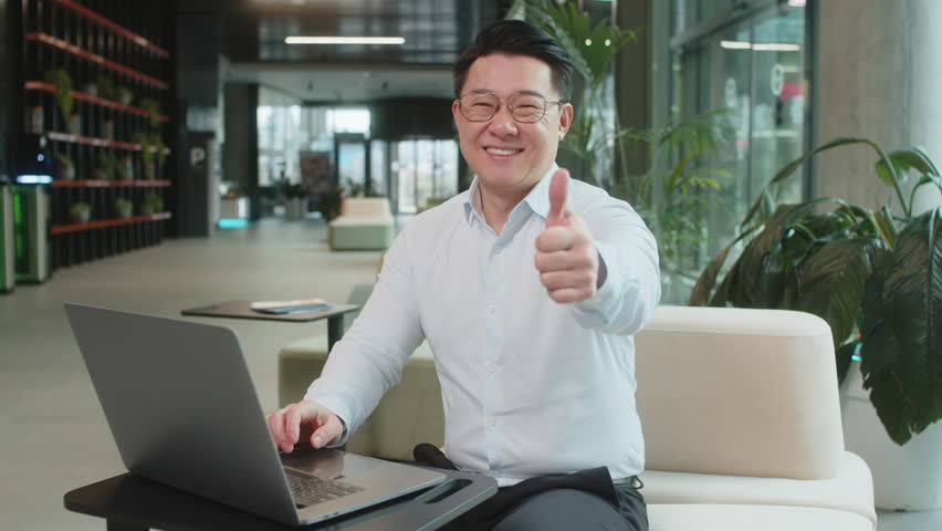Like. Happy young businessman guy working on laptop looking approvingly at camera showing thumbs up, like sign positive something good, positive feedback sitting in modern office lobby. Businesspeople