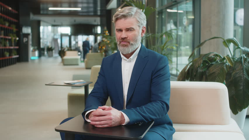 Dislike. Upset businessman guy in suit sitting in office lobby showing thumb down sign gesture, expressing discontent, disapproval, dissatisfied bad work. Displeased serious mature man in hotel hall.