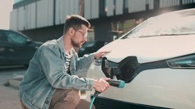 Focused young Caucasian male in denim jacket plugging charging cable into electric vehicle Glowing holographic diagrams floating nearby. Digital interface displaying battery levels. AI integration. - Powered by Shutterstock - Get 15% off with code: PIKWIZARD15