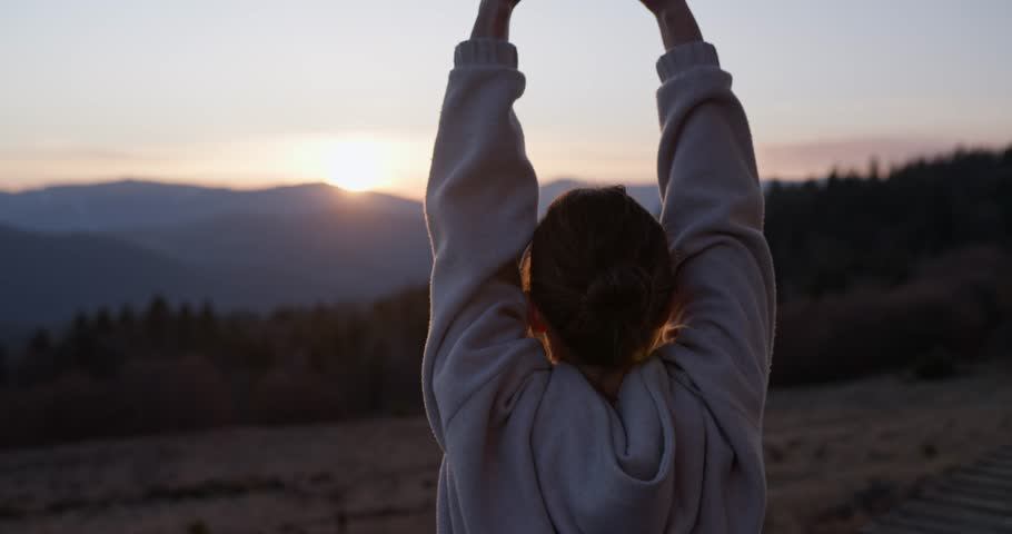 Back view of girl doing sports and stretching while watching sunset on porch of her country house among forest and mountains