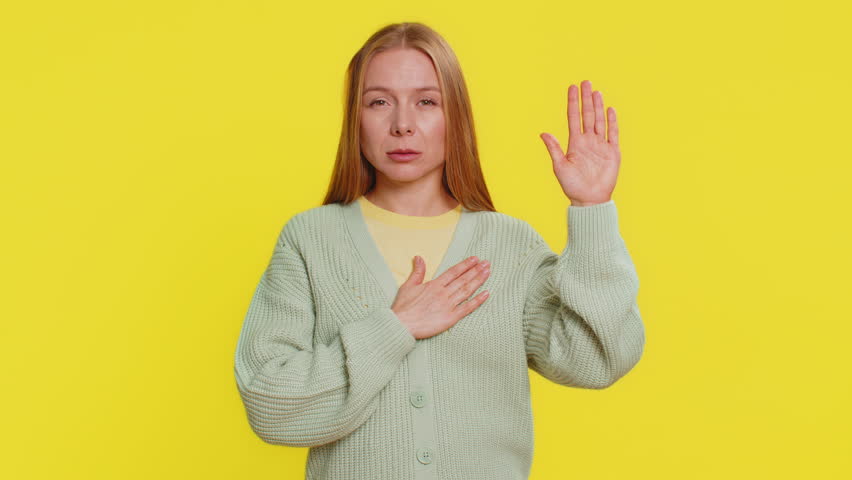 I swear to be honest. Sincere responsible Caucasian young woman 40 years old raising hand to take oath, promising to be honest, to tell truth, keeping hand on chest, justice. Girl on yellow background