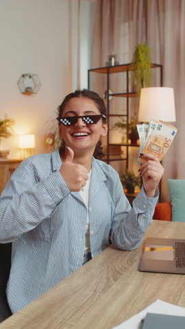 Young businesswoman freelancer putting on party glasses holding fan of cash money euro banknotes showing thumbs up, celebrate, success career, online income, wealth at home office desk. Vertical.