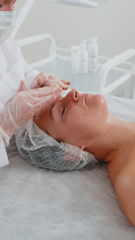 Cosmetologist applying special moisturizing medical cream, scrub, peeling face of pretty Caucasian woman. Facial skin care anti aging cosmetic massage procedure in a beauty salon. Beauty treatment