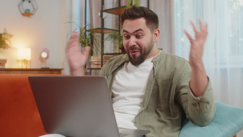 Surprised Caucasian man with brown hair using laptop, receive mail good news message, wow shocked by sudden victory celebrate lottery jackpot win purchases online shopping at home on sofa. Lifestyles.