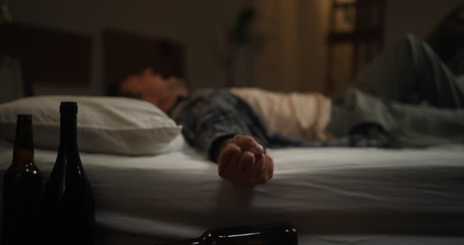 Man in checkered shirt lying on white mattress screaming and trying to rest after despair and drinking alcohol with empty bottles in room. A man on a drinking binge