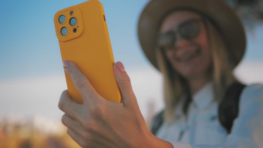 Smiling young woman traveler wear hat and backpack capturing vacation moment, taking selfie with bright yellow smartphone against blurred sky background.Travel, blogging, weekend, relax and lifestyle