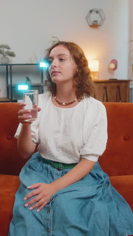 Satisfied young Caucasian woman sitting at home on sofa couch leading healthy lifestyle, drinking a glass of pure water, smiling. Drink daily norm of water. Girl smiling at camera at home. Vertical