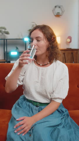 Satisfied young Caucasian woman sitting at home on sofa couch leading healthy lifestyle, drinking a glass of pure water, smiling. Drink daily norm of water. Girl smiling at camera at home. Vertical