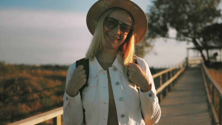 Blonde traveler walking wooden boardwalk at golden sunset, backpack-wearing, sunhat-clad, waving cheerfully amid scenic landscape.Europe travel. Lifestyle, vacation, tourism, nature, active life
