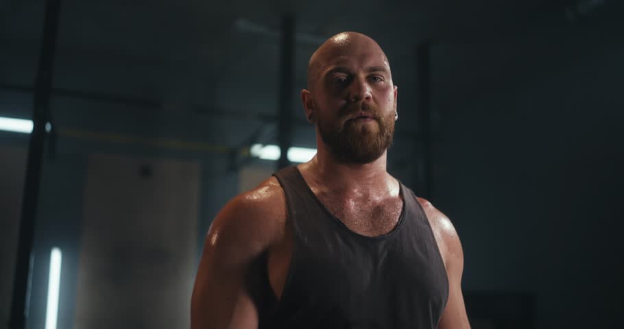 Portrait of a strong muscular man with a beard and shaved head who wipes his forehead from sweat in a gray gym