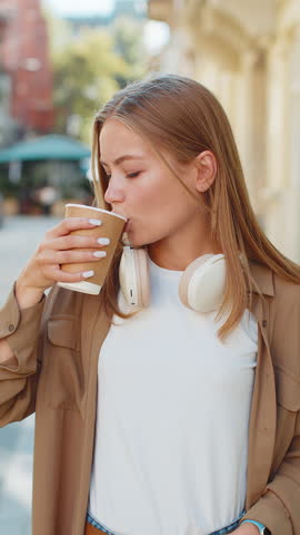 Happy Caucasian woman enjoying morning coffee hot drink and smiling. Relaxing, taking a break. Young girl teenager walking on urban city center street, drinking coffee to go. Town lifestyles outside.