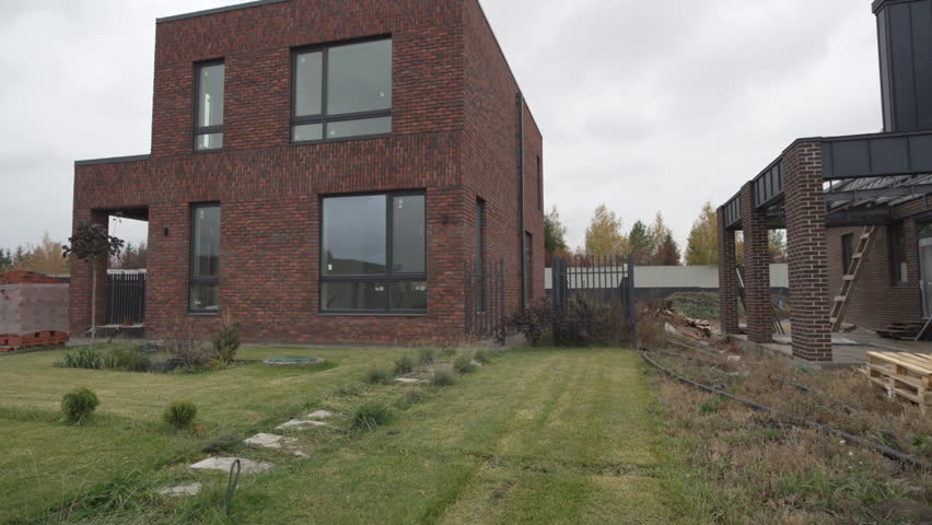 Two-story brick house with modern minimalist design and panoramic windows. Grass lawn with stone pathway along with young bushes in yard