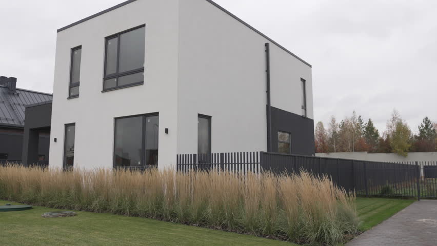 Two-story house features minimalist design with contrasting facade. Large windows metal fence and lawn enhance contemporary aesthetic