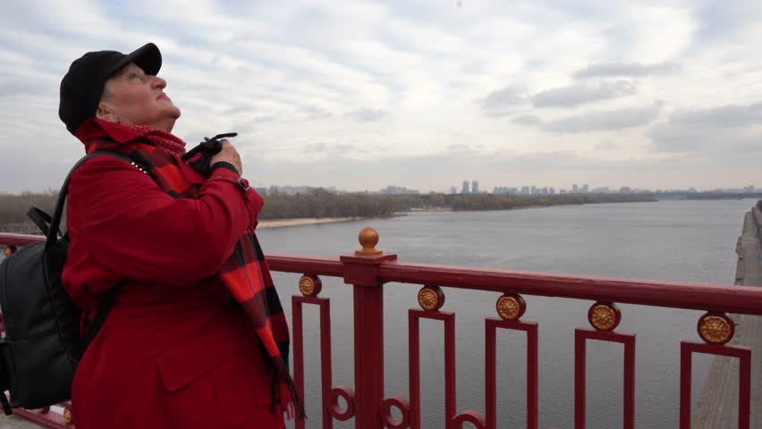 Elderly woman stands on a bridge over a river. A grandmother walks outside in cold weather. She is wearing a hat and coat. Outdoor