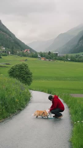 Person in red jacket kneels to pet golden retriever on a serene pathway in a green valley surrounded by mountains