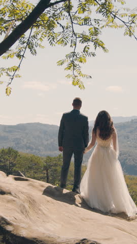 A happy couple of brides stand on the mountain with their backs to the camera and look into the distance at the mountains covered with trees. Looking into the future of a young couple in love.