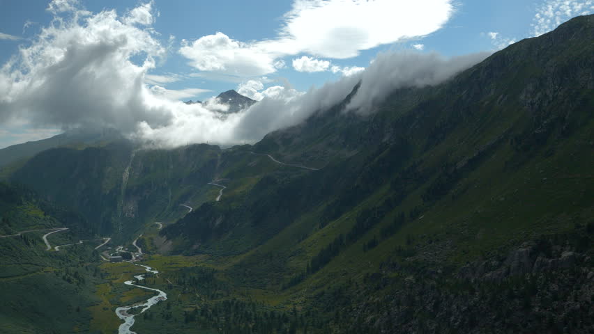 TIME LAPSE: Dramatic alpine road winds up through green mountain slopes beneath thick, sweeping clouds. Raw beauty of the Swiss Alps, with serpentine road leading through misty ridgelines and valleys.