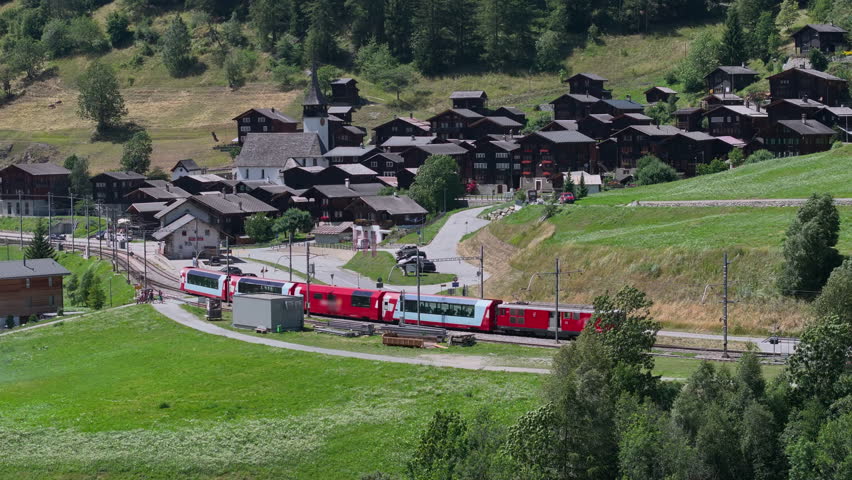 AERIAL: Red train passing through charming Swiss mountain village with traditional wooden alpine houses and white steepled church. Traveling through the picturesque green alpine countryside by rail.