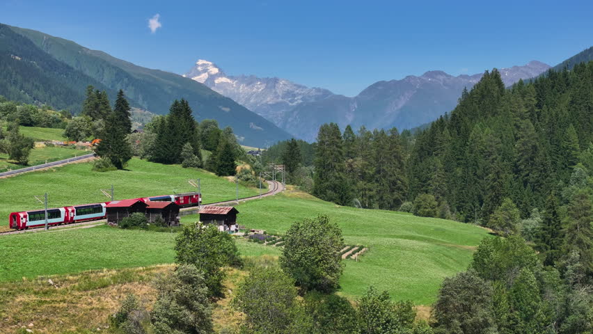 AERIAL: Train travels through a picturesque Swiss alpine valley, passing through pine forests, rustic villages and with views of majestic mountains. Scenic route through the mountainous countryside.