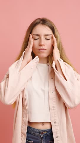 Young woman experiencing a headache, touching her temples and forehead with her hands, expressing discomfort and pain on a pink background