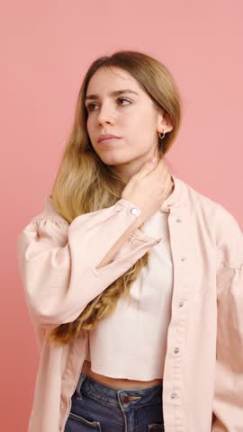 Studio shot of a young woman experiencing neck pain and stiffness, massaging the affected area for relief on a pink background