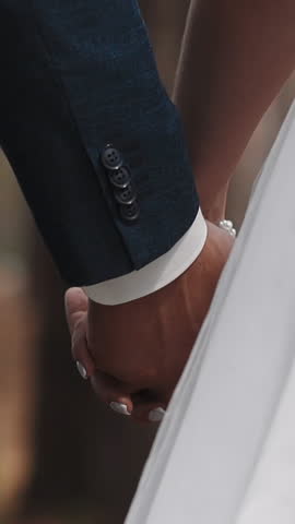 Newlywed wife and husband walk joining hands with interlaced fingers at wedding ceremony across park closeup slow motion. Young man and woman couple in love