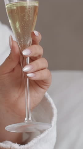 Young woman wearing white bathrobe holds glass of champagne sitting on bed in hotel room close view. Lazy morning at vip-resort. Vacation and relax
