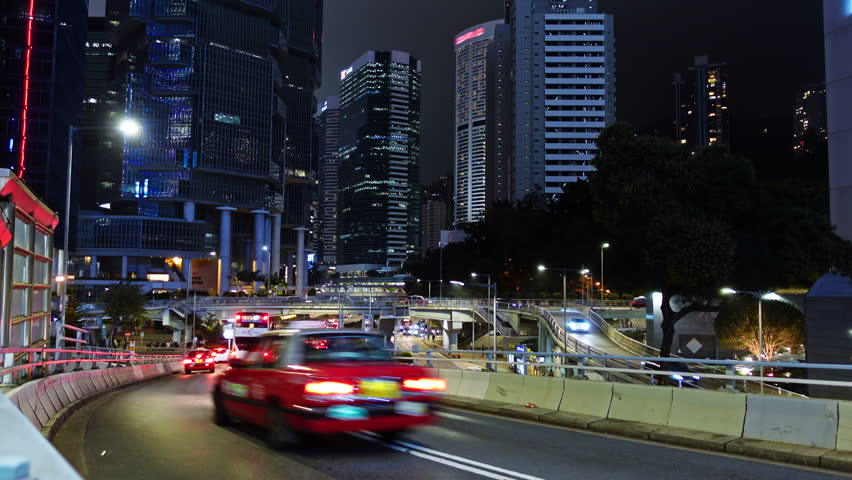 Nighttime urban traffic flows along winding roads in Hong Kong. Brightly lit skyscrapers tower in the background, showcasing the city