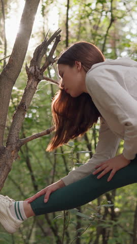 Young lady stretches for warming up in forest. Woman prepares before going for run in summer morning surrounded by trees. Active routine for well-being