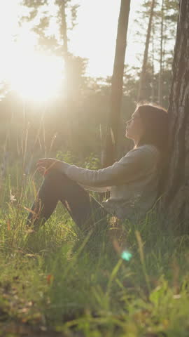 Lady leans on tree trunk bark in forest at sunset. Young woman rests in fresh air thinking about mental health improvement. Coping with personal problems in nature