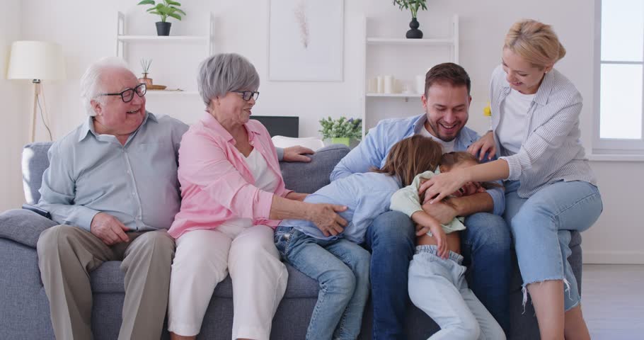 Big happy family gathering on sofa with parents and grandparents playfully tickling children. Joyful multi generation relatives having fun at home, teasing their lovely kids, enjoying time together.