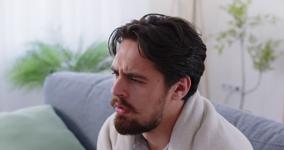 Sick man stays at home and drinks medicine. Unhappy young man, sitting on the sofa in a warm blanket, coughs, blows his nose in a tissue, and takes bitter medicine with water. Concept of cold and flu 