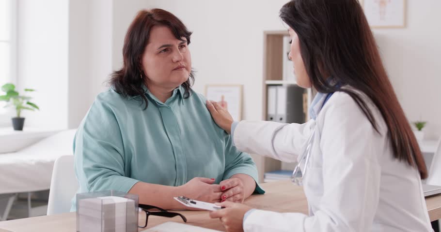 Friendly doctor talks to worried anxious fat chubby ill woman patient with cardiovascular heart disease, gives professional therapy recommendation writes prescription on clipboard, supports, cheers up