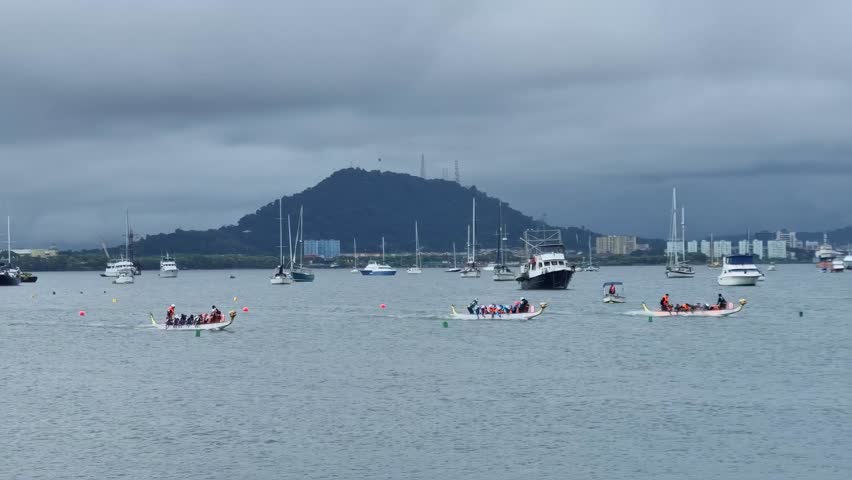 Dragon boat race in bay with cloudy sky and mountain in background, surrounded by sailboats and yachts.