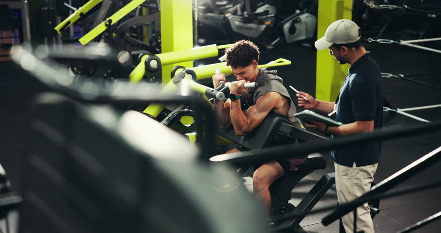 Men, workout or bicep curl with personal trainer at gym for strength training or arm exercise on machine. Male people, fitness instructor or coach with tablet for physical routine or health checklist